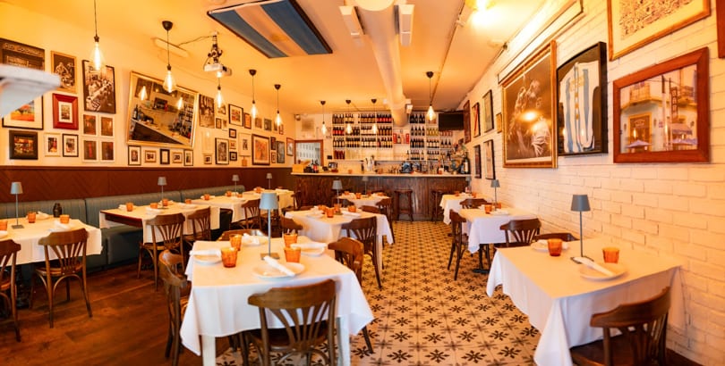 Cozy restaurant interior with white tablecloths, wooden chairs, a bar, and framed art on the walls.
