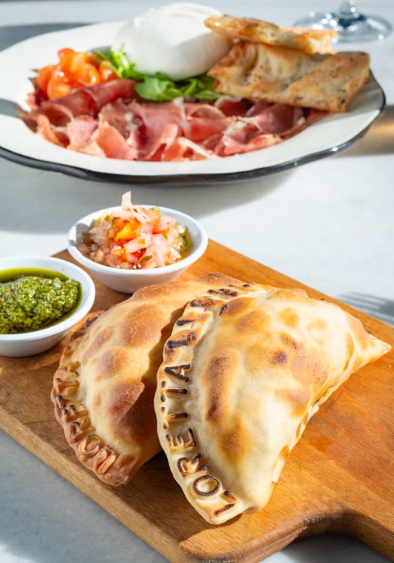 Two empanadas with dipping sauces on a wooden board, and a burrata salad in the background.