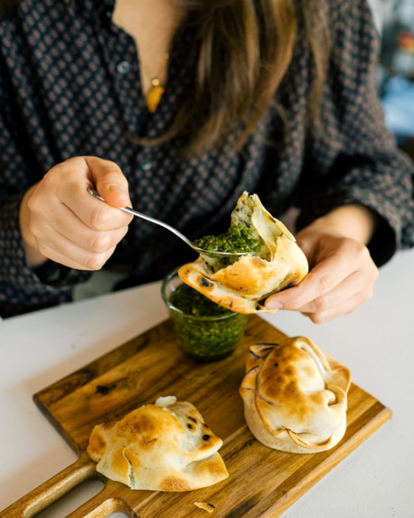 Person spoons green chimichurri sauce into a golden empanada on a wooden board.