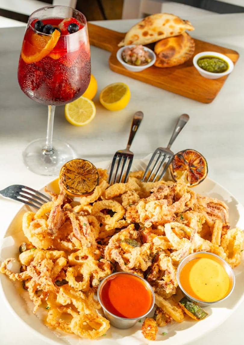 Fried calamari with sauces, sangria, empanadas, and lemons on a white table.
