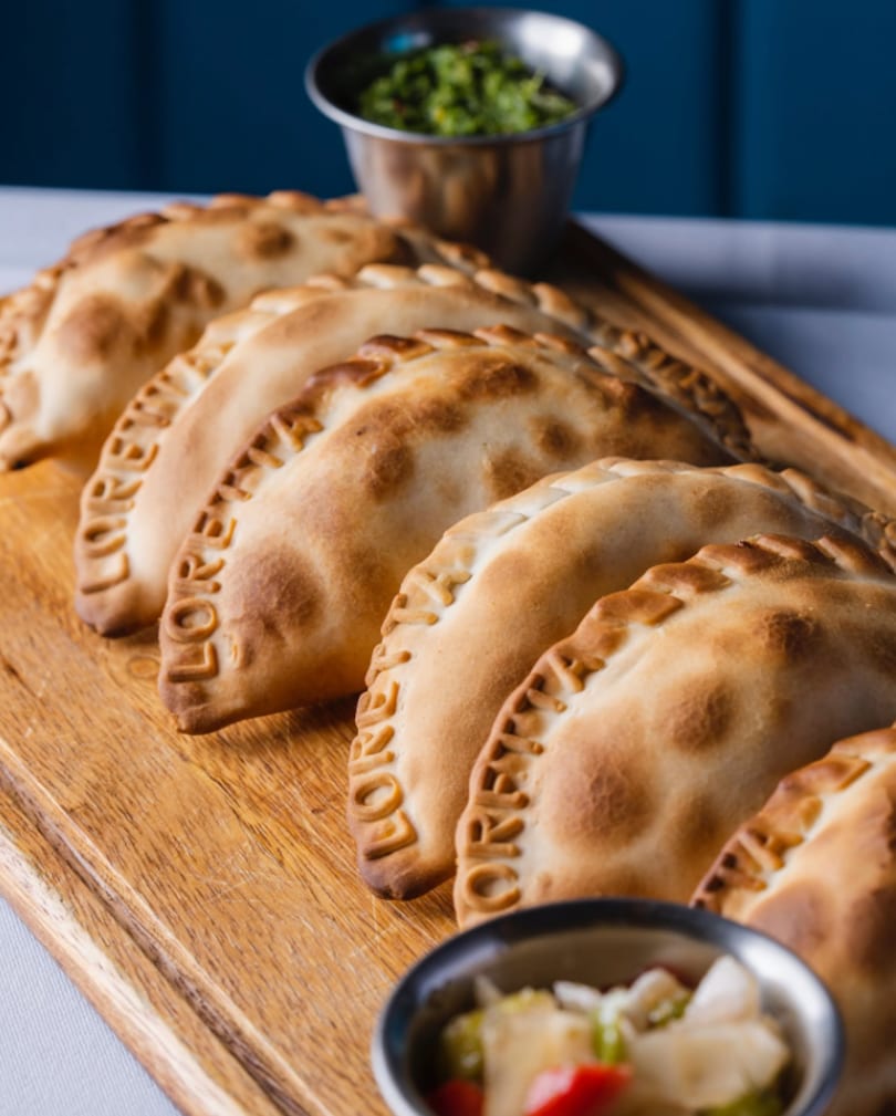 Row of golden-brown empanadas with "LORENA" embossed on them, served on a wooden board with sauces.