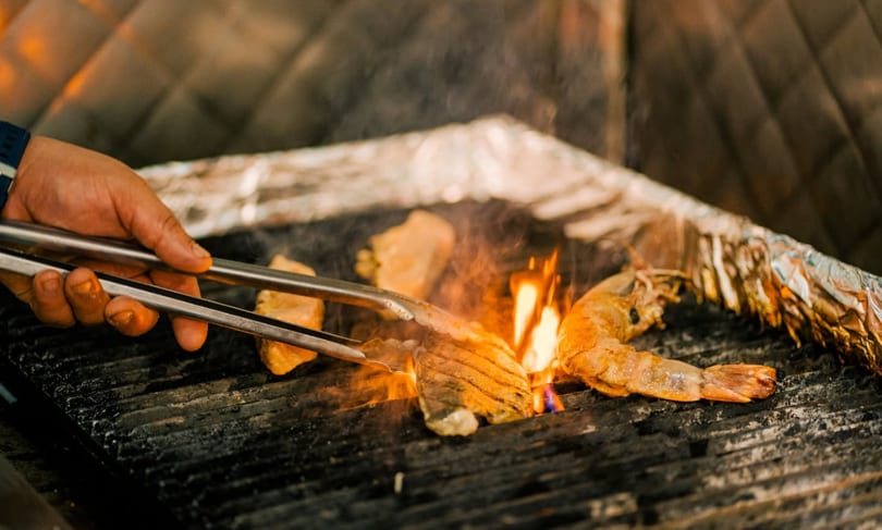 A hand uses tongs to flip shrimp and fish fillets cooking on a fiery grill.